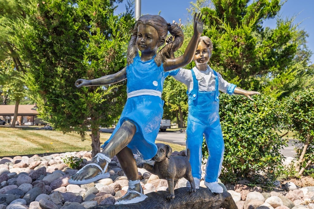 A statue of two children in blue overalls and white shoes is standing on a rocky surface with trees in the background.