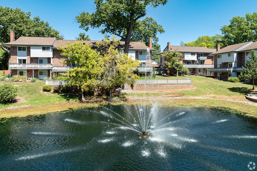 A fountain in the middle of a pond in front of apartment buildings.