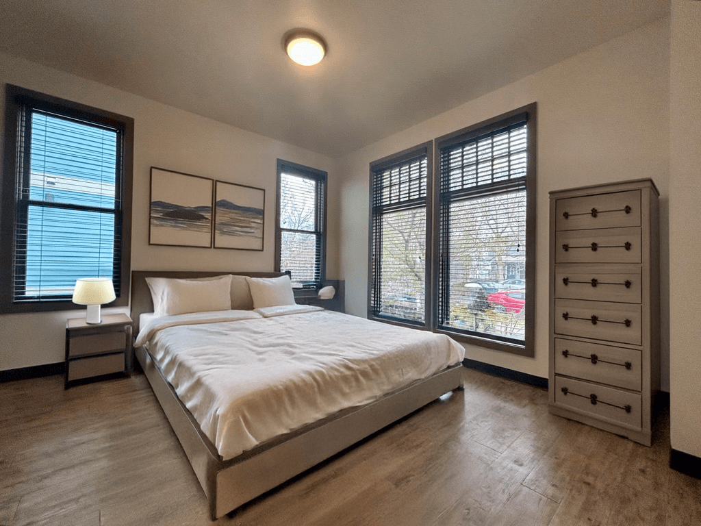 A bedroom with a large bed, a nightstand with a lamp, and a dresser with a mirror.