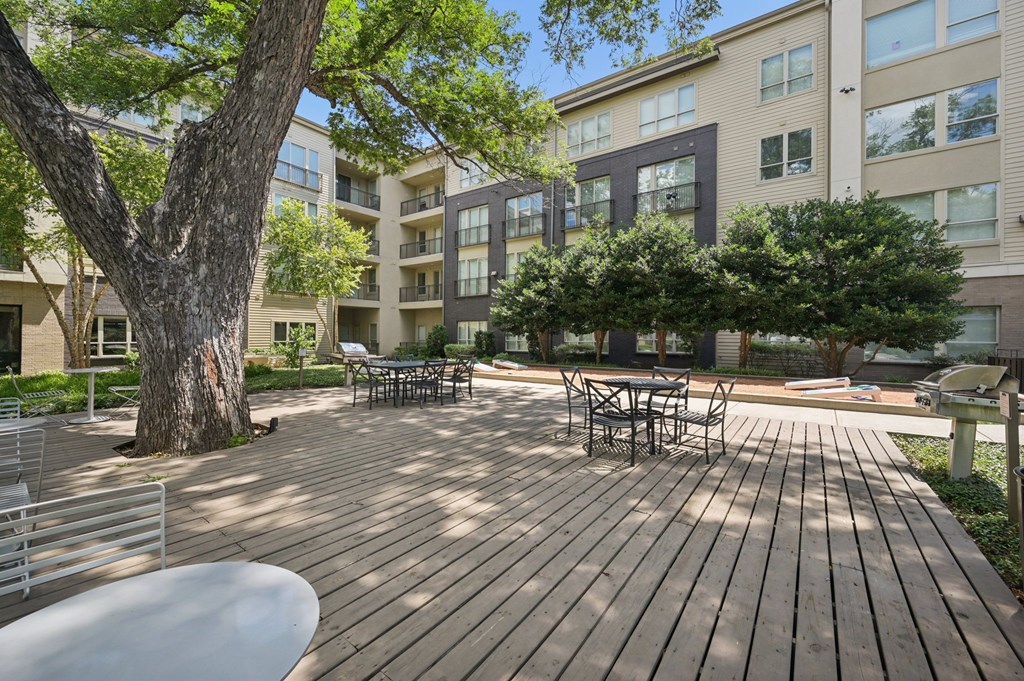 A wooden deck with a table and chairs is surrounded by trees and buildings at Everra Midtown Park Apartments, Dallas 75231