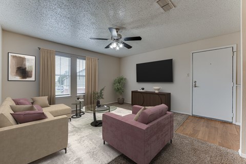 A living room with a beige couch, a pink ottoman, a black TV, and a ceiling fan.