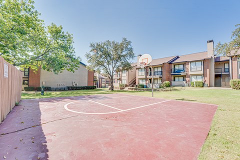 A basketball court is located in the middle of a grassy area in front of apartment buildings.