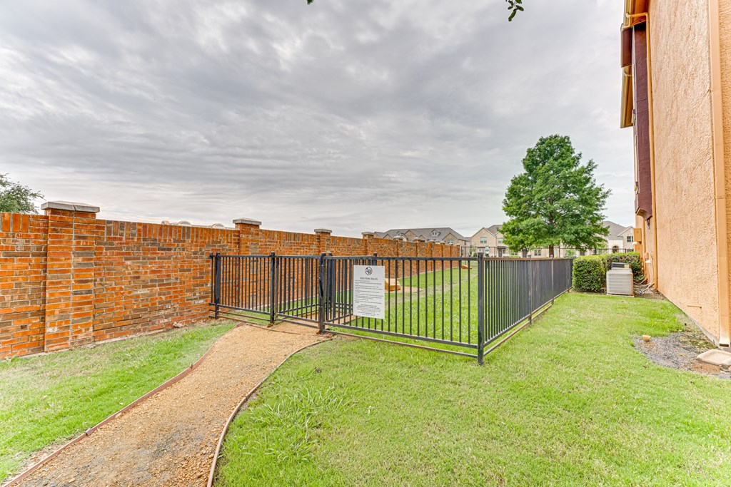 A black metal fence surrounds a grassy area with a brick wall on one side and a dirt path leading to a gate at Limestone Ranch Apartments, Lewisville 75067