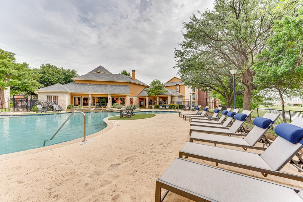 A row of lounge chairs are set up on a sandy area in front of a pool and a building at Limestone Ranch Apartments, Lewisville, Texas