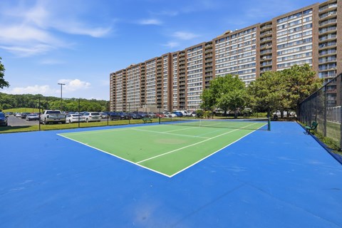 A tennis court with a blue surface and white lines is surrounded by apartment buildings.