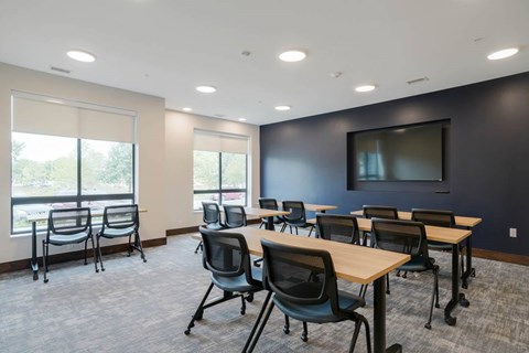 Conference room with light wood tables, mesh chairs, large windows with shades, and wall-mounted TV, offering natural light and open layout.