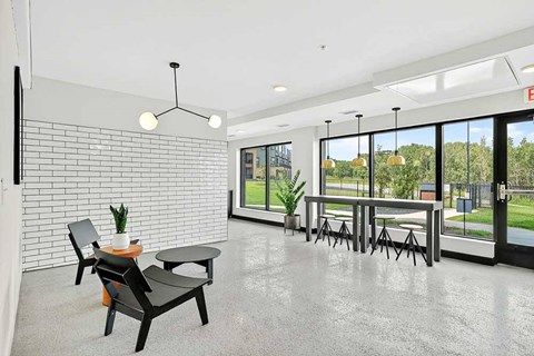 Bright apartment community lounge with polished concrete floors, modern seating, white tile accent wall, and large windows with views of green outdoor space.