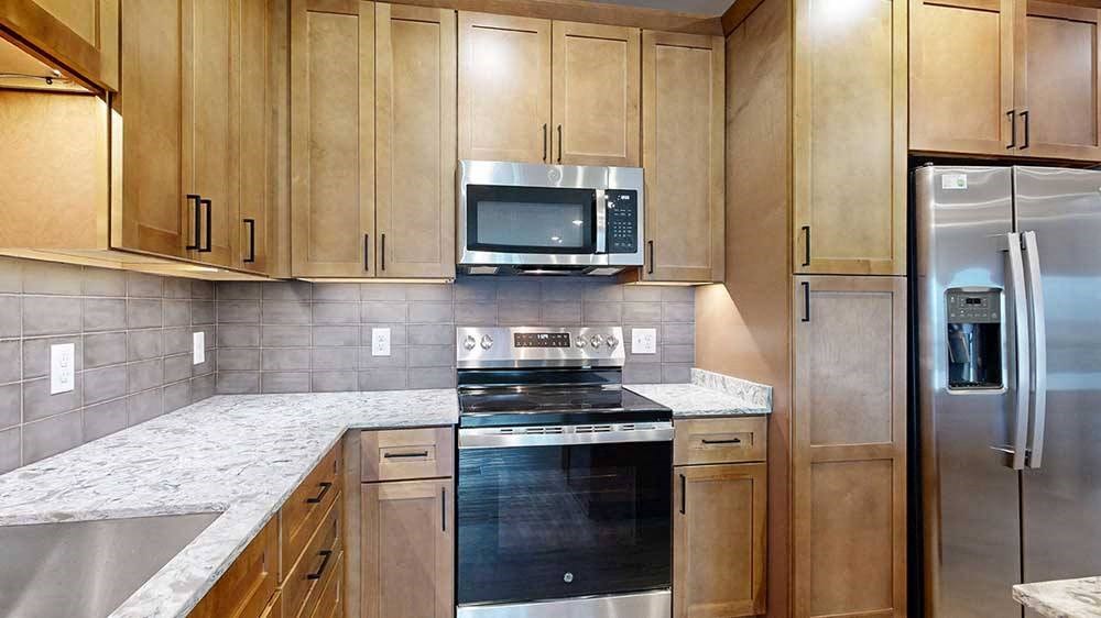 a kitchen with light brown wooden cabinets, quartz countertops, a grey tile backsplash, and stainless steel appliances