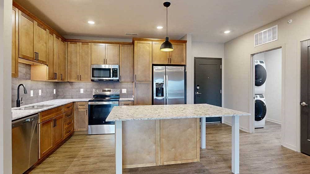 a kitchen with a large table island, light brown wooden cabinets, quartz countertops, and stainless steel appliances