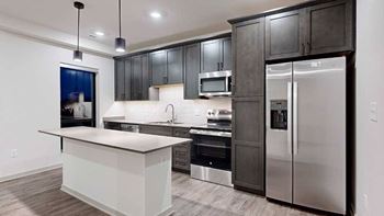 a modern kitchen with dark wood cabinets, light quartz countertops, a narrow central island, a tile backsplash, and stainless steel appliances