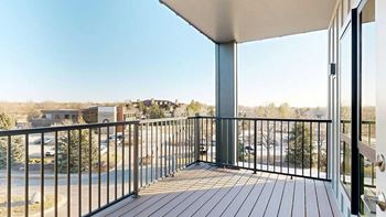 A balcony with a black railing and wooden floor overlooks a parking lot and buildings.