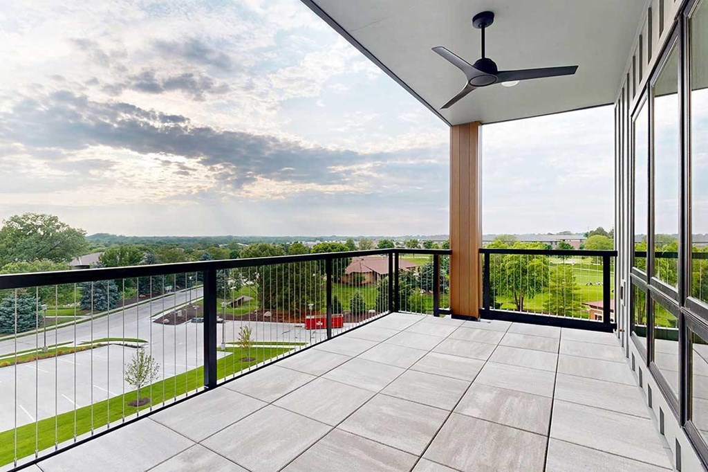 An oversized balcony with a metal and wire railing, stone tile flooring, a ceiling fan, and a corner view of the neighborhood.