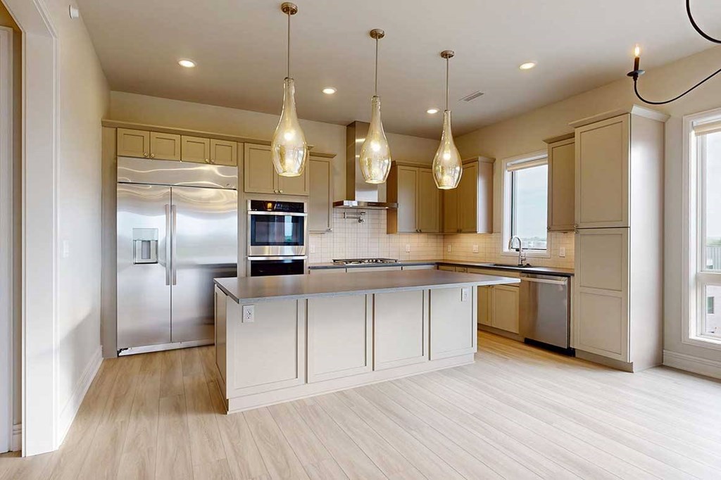 A gourmet kitchen with an oversized island, taupe wooden cabinetry, stainless steel commercial appliances, quartz countertops, a range hood, a tile backsplash, and pendant lighting.