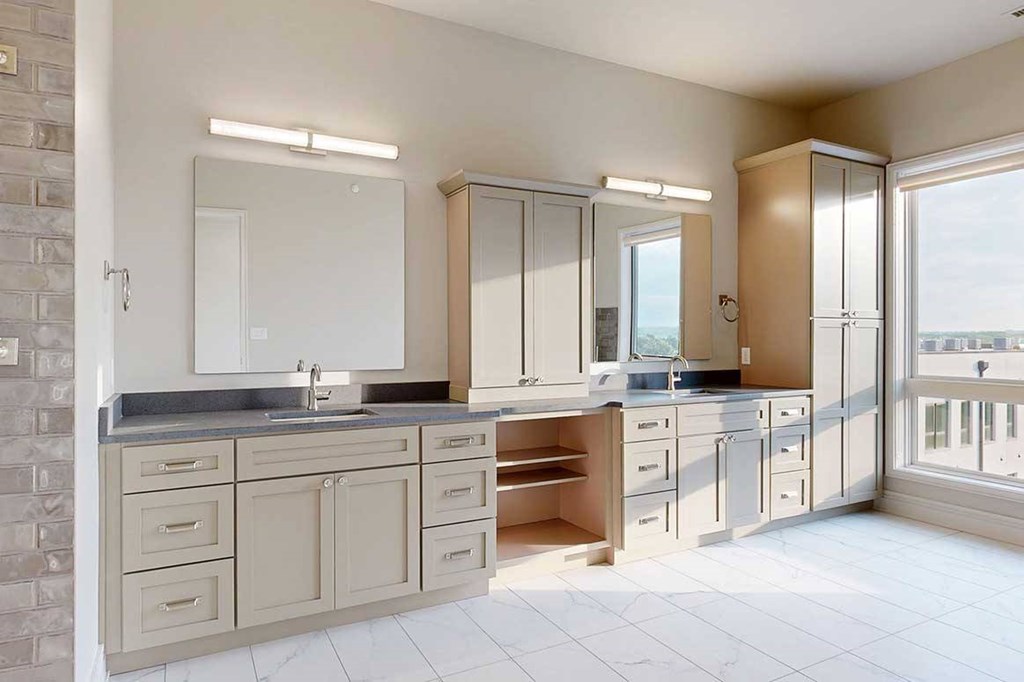A spacious ensuite bathroom featuring a quartz double vanity with taupe cabinetry, a tall storage cabinet, and tiled flooring.