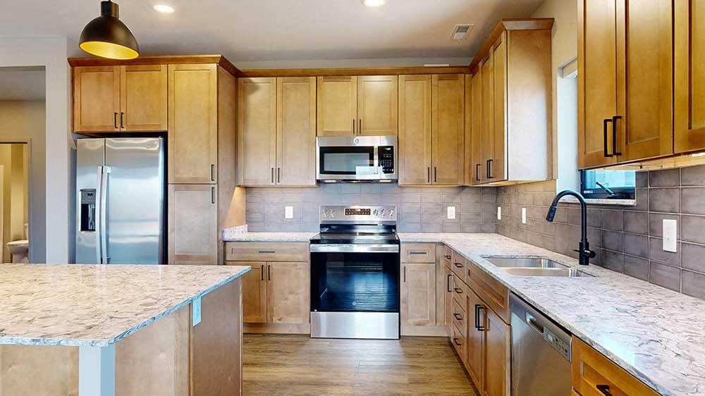 a kitchen with light brown wooden cabinets, stainless steel appliances, a tile backsplash, a large central island, and quartz countertops