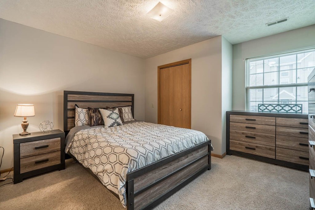 A bedroom with a large bed, a nightstand with a lamp, and a nook with a large window