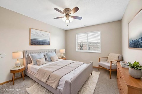 Virtually staged bedroom with a large bed, an accent chair, a ceiling fan, and a window bringing natural light into the space.