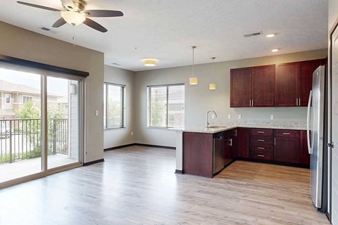 An open concept living room with a sliding glass door to a balcony, kitchen with wooden cabinets, and a dining area in the background.