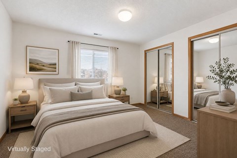 Bright, virtually staged apartment bedroom with neutral carpeting, mirrored closet doors, wood trim, and a large window with natural light.