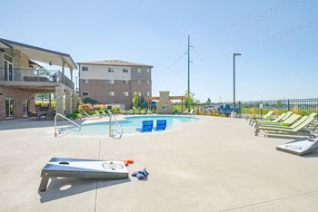 a pool with green lounge chairs and a cornhole game in front of a building