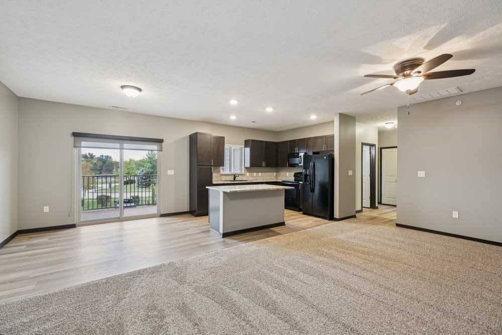 A spacious, carpeted living room open to a modern kitchen with hardwood flooring and dark wooden cabients.
