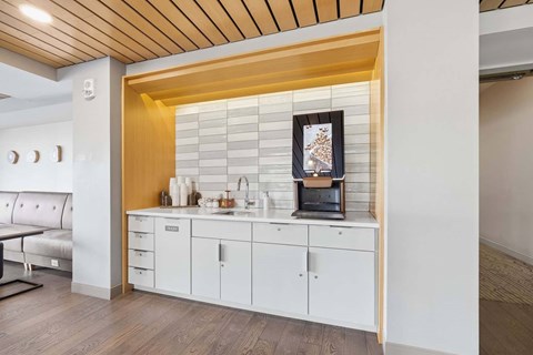 Community coffee station with a beverage machine, sink, white cabinets, tiled backsplash, and wood accents.