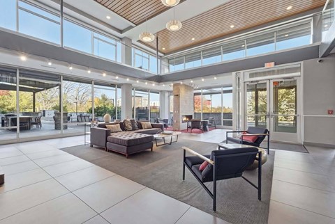 Community lounge with a sectional sofa, armchairs, fireplace, double-height windows, and views of an outdoor patio.