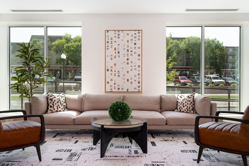 Community lounging space with large beige couch in between two large windows. Two chair sit opposite of the couch with a small coffee table at the center.