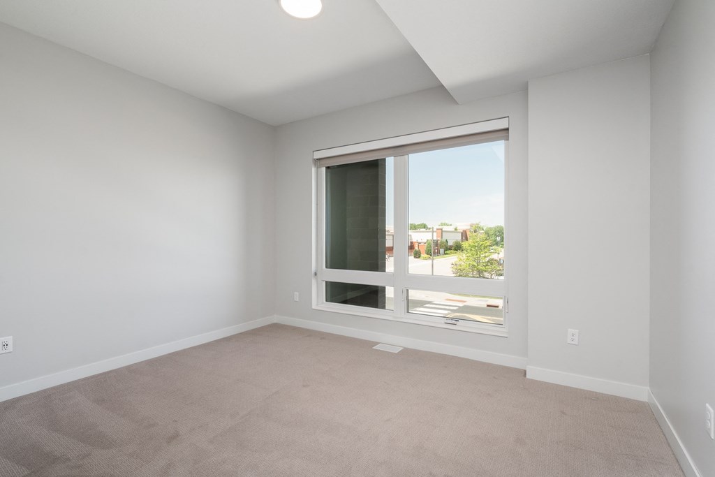 Second bedroom with large floor to ceiling window, beige carpet, and white walls.