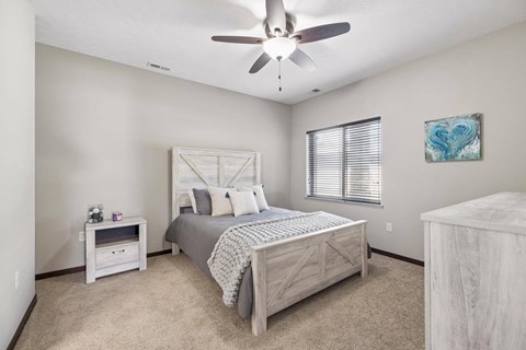 Staged apartment bedroom with ceiling fan, carpet, wood-look bed frame, dresser, and window with blinds.