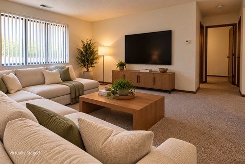 Virtually staged apartment living room with a spacious sectional, large window, carpeted flooring, and an open hallway.