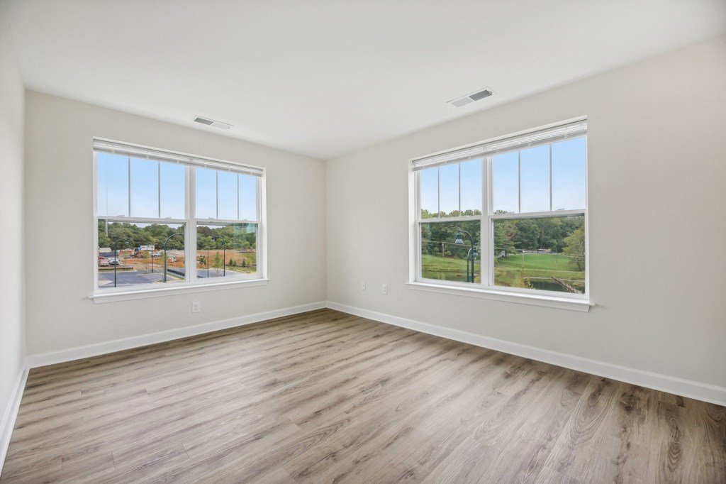A room with two windows overlooking a street and a green field.
