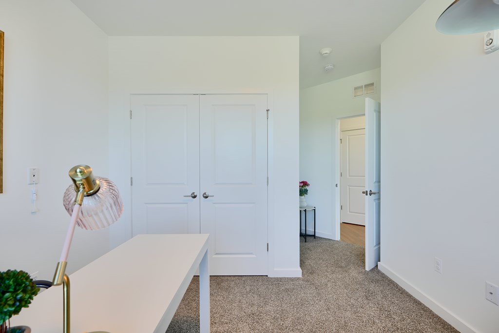 Now leasing senior 1-bedrooms in Baltimore, MD. A white table with a gold lamp stands in a room with white walls and a carpeted floor.