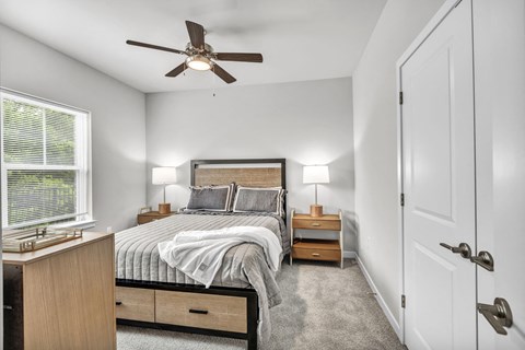 Bright bedroom with a modern bed frame and layered bedding, complemented by wood nightstands, table lamps, and a ceiling fan centered above.