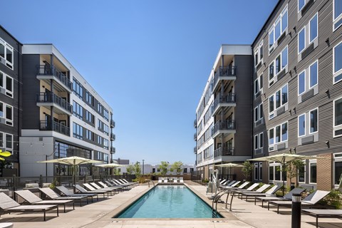 A swimming pool surrounded by lounge chairs and umbrellas in front of apartment buildings.
