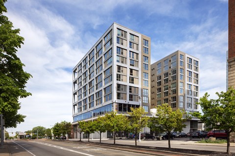 A modern multi-story building with a glass facade is situated on a street corner with trees and other buildings in the background.