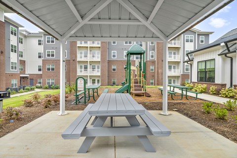 Community courtyard view from a covered pavilion, featuring picnic seating in the foreground and a children’s play area surrounded by walkways and greenery.