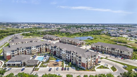 Aerial view of a modern apartment community featuring landscaped grounds, a resort-style pool, and surrounding green space.