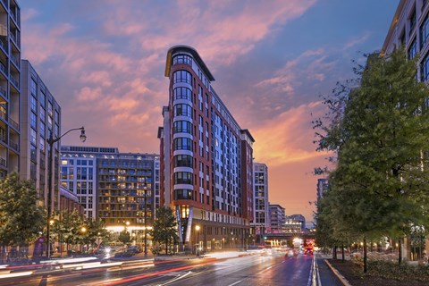 A city street in the at dusk with cars passing by and buildings on either side.