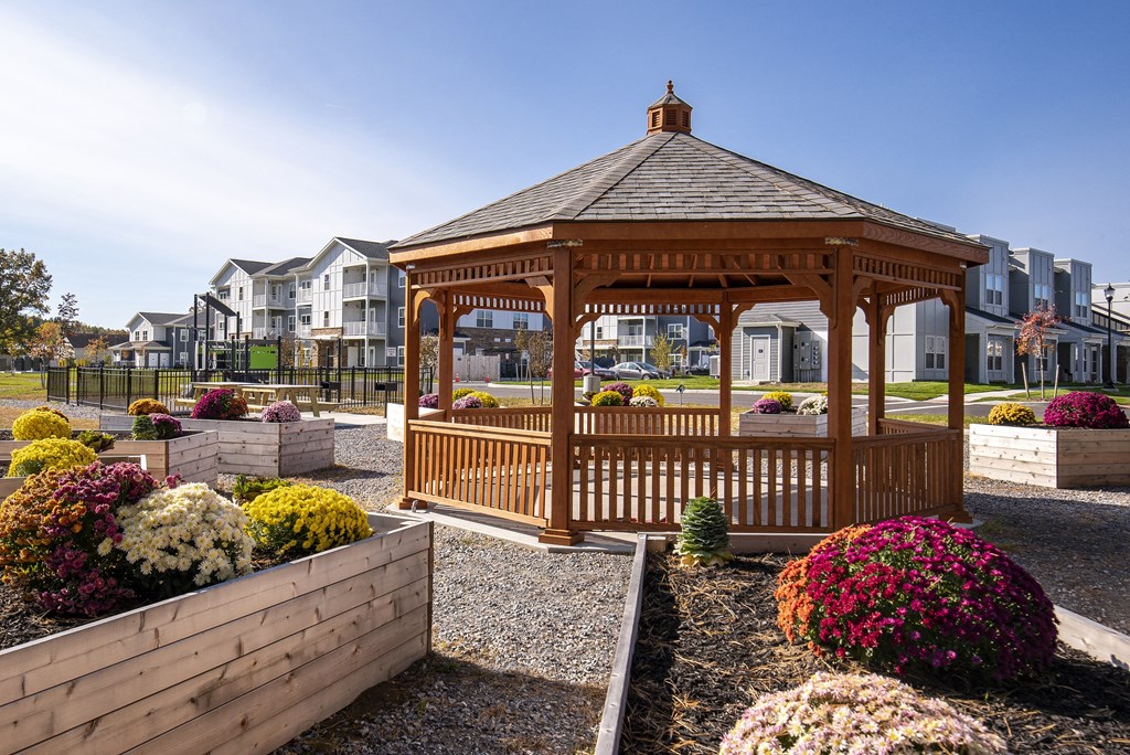 A wooden gazebo is surrounded by a gravel area and flower beds.