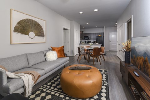 A living room with a grey couch, a round orange ottoman and a painting on the wall.