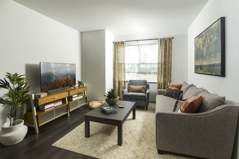 A living room with a grey couch, a wooden coffee table, a painting on the wall, and a large window with curtains.