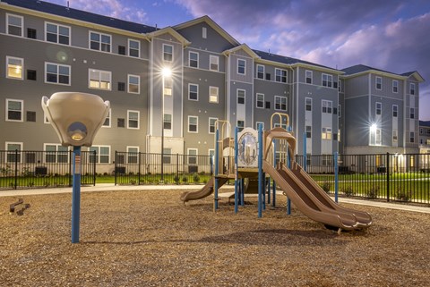 Community playground with slides and climbing structures set within a landscaped outdoor area, with apartment buildings in the background.