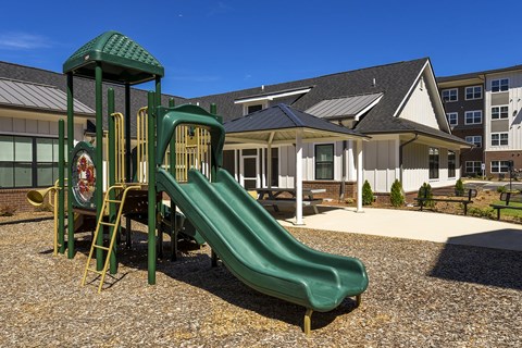 Outdoor playground featuring multi-level climbing equipment and slides, positioned next to a shaded seating pavilion and landscaped grounds.