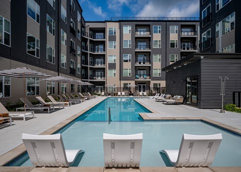 A swimming pool surrounded by white chairs and umbrellas in front of a multi-story apartment building.