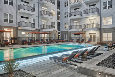 A swimming pool surrounded by lounge chairs and umbrellas in front of apartment buildings.