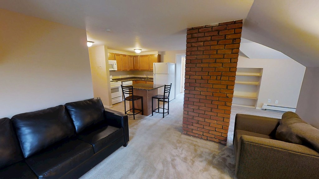 A black leather couch sits in a living room with a brick pillar and a kitchen in the background.