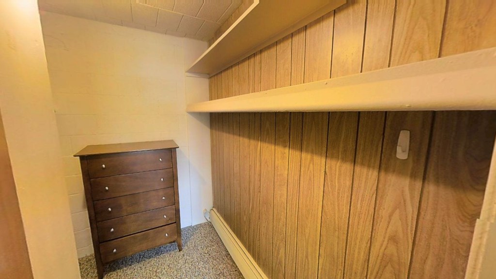 A wooden dresser sits in a room with a white wall and a wooden shelf above it.