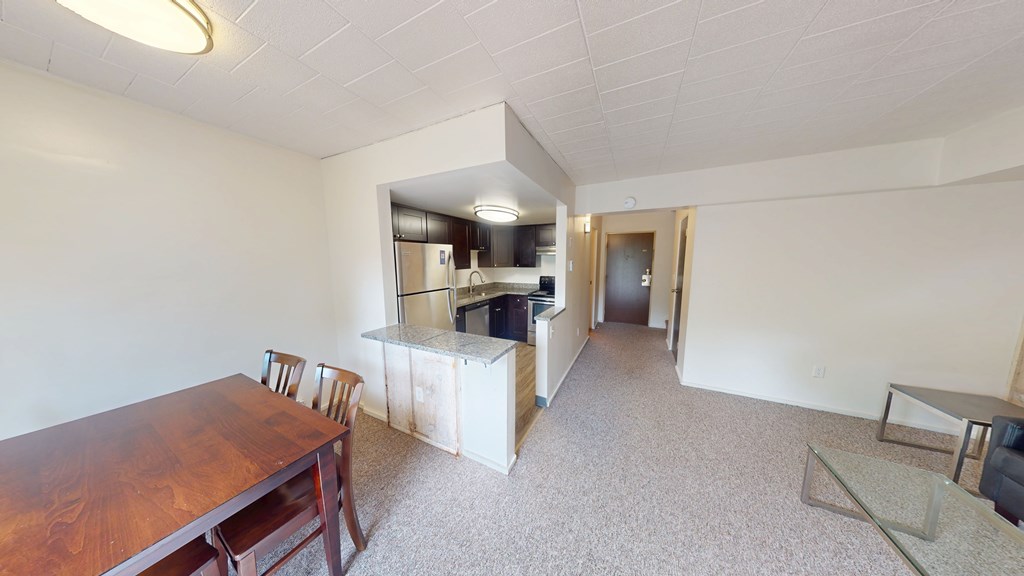 A room with a wooden table and chairs in the foreground and a kitchen area in the background.