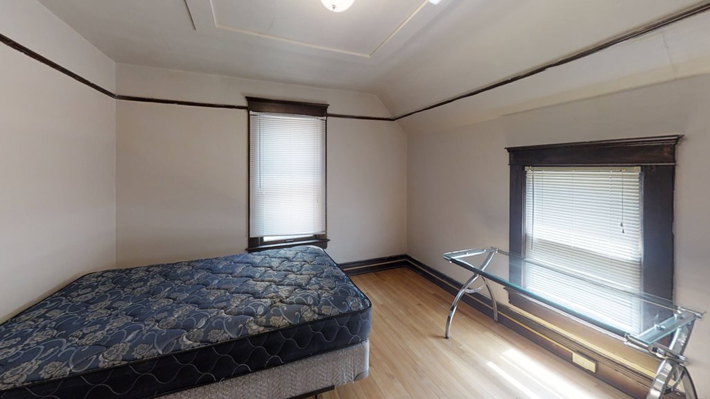 A bedroom with a bed, a glass table, and a window with blinds.